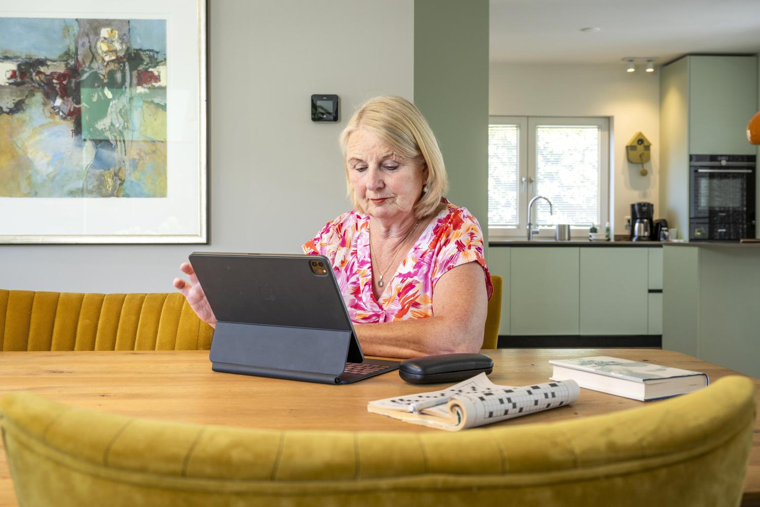Mevrouw die aan de keukentafel zit en op een Ipad aan het kijken is. 